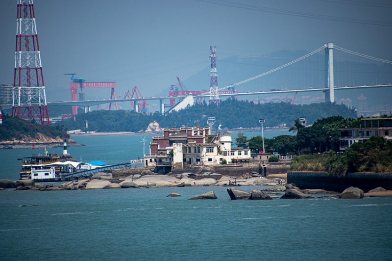 In the foreground is an old colonial mansion, in the background a modern suspension bridge and portions of the modern port operation in Xiamen. The Port of Xiamen is an important deep water port located on Xiamen Island, the adjacent mainland coast, and along the estuary of the Jiulongjiang River in southern Fujian, China. It is one of the trunk line ports in the Asia-Pacific region. It is ranked the 8th-largest container port in China and ranks 17th in the world. It is the 4th port in China with the capacity to handle 6th-generation large container vessels.